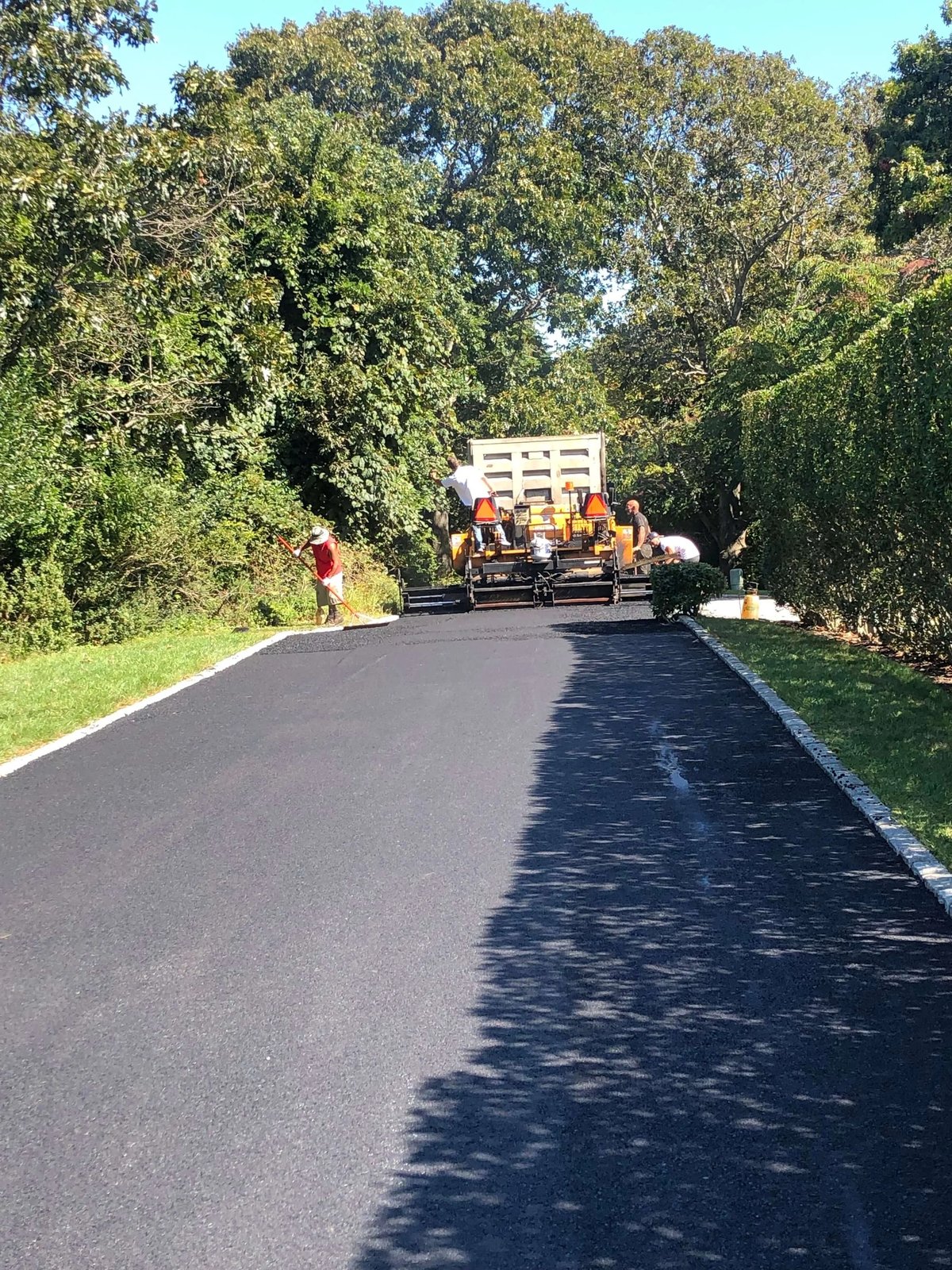 Long private road being resurfaced with fresh asphalt beside a granite curb and hedgerow